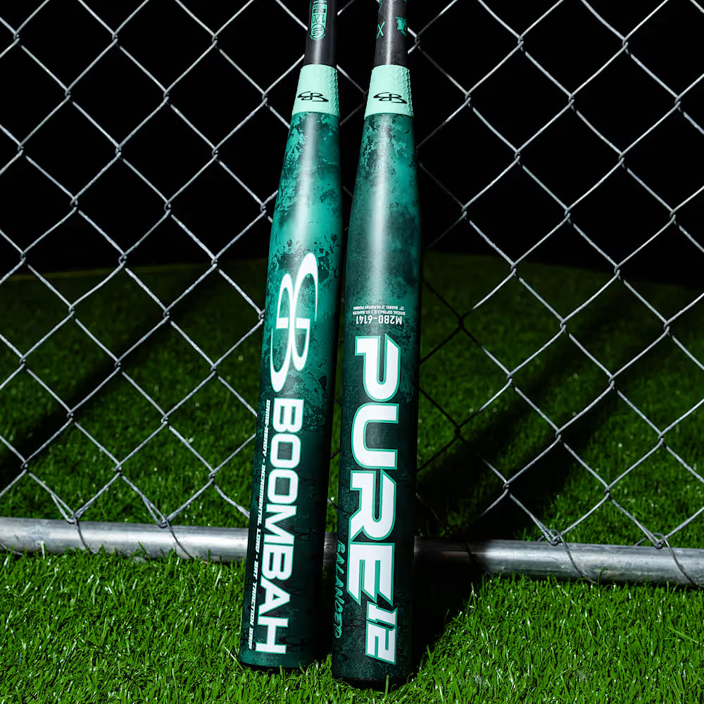Two green and black BoombahPure Pure12 Teal Control 12" Balanced Slowpitch Softball Bats, model M2B0-6141 TL, lean against a chain-link fence on artificial turf at night, displaying Boombah branding and X25 Barrel Technology.