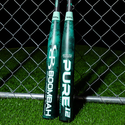 Two green and black BoombahPure Pure12 Teal Control 12" Balanced Slowpitch Softball Bats, model M2B0-6141 TL, lean against a chain-link fence on artificial turf at night, displaying Boombah branding and X25 Barrel Technology.