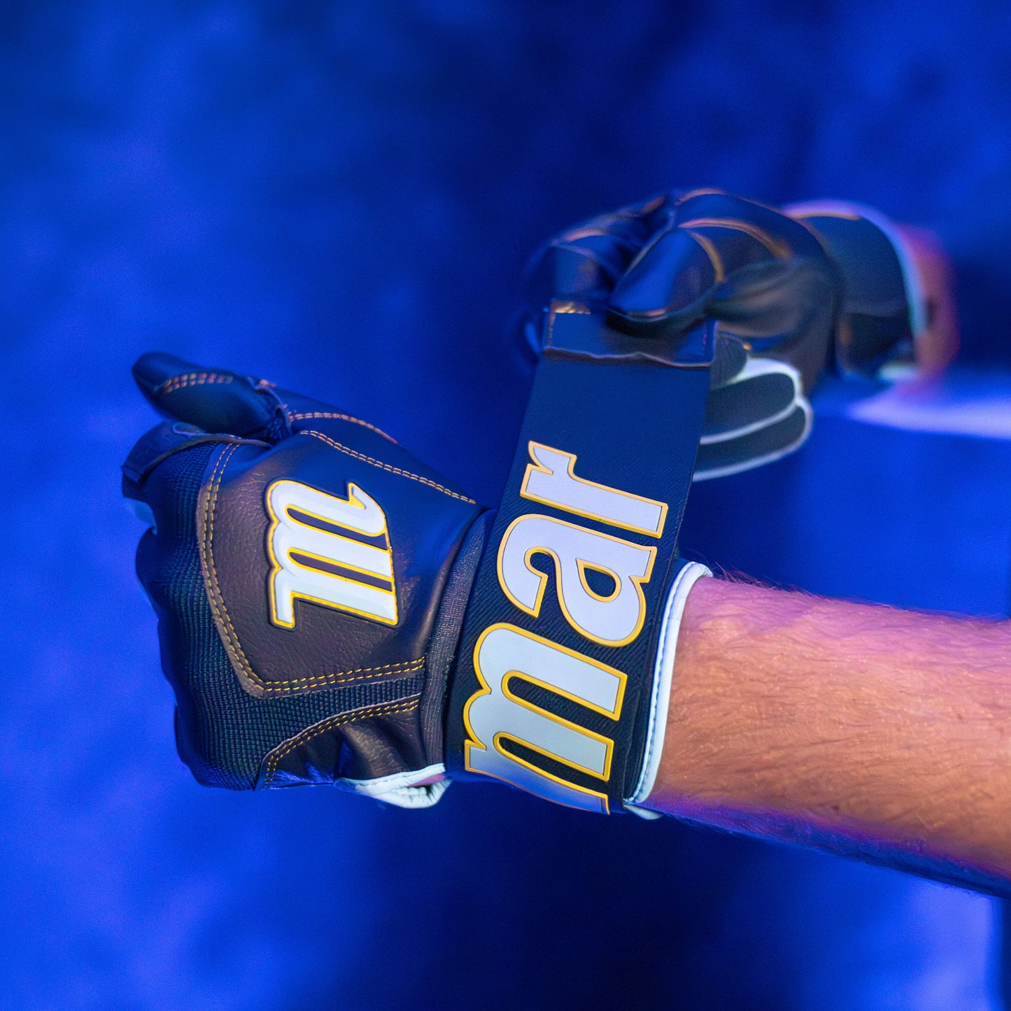 A person wearing durable Marucci Blacksmith V2 Full Wrap Youth Batting Gloves fastens one glove with a large strap displaying “Marucci” in bold yellow letters under blue lighting.