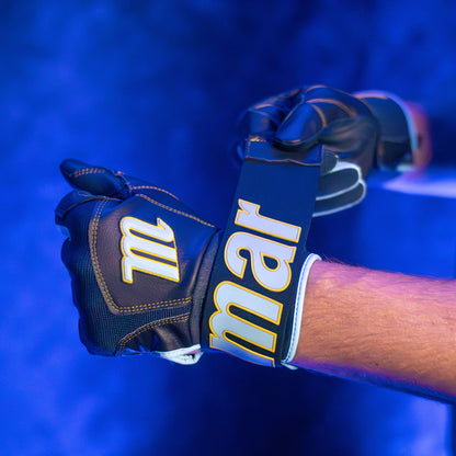 A person wearing durable Marucci Blacksmith V2 Full Wrap Youth Batting Gloves fastens one glove with a large strap displaying “Marucci” in bold yellow letters under blue lighting.