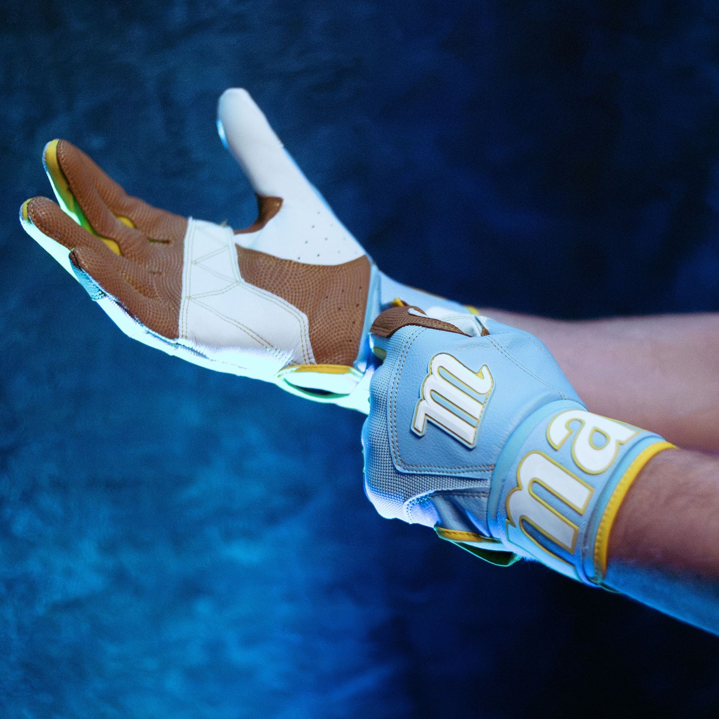 A person puts on Marucci Blacksmith V2 Full Wrap Adult Batting Gloves (MBG2BKSM) in light blue and white with yellow accents, featuring reinforced padding for extra protection, against a dark blue textured background.