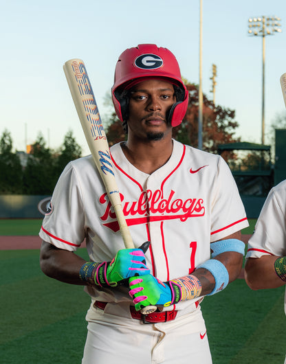 A baseball player in a white Bulldogs uniform with a red helmet grips the Marucci CATX RCKLESS Rewind Hybrid (-3) BBCOR Baseball Bat (MCBCRHRC), ready to swing on the field under stadium lights, wearing colorful gloves and wristbands.