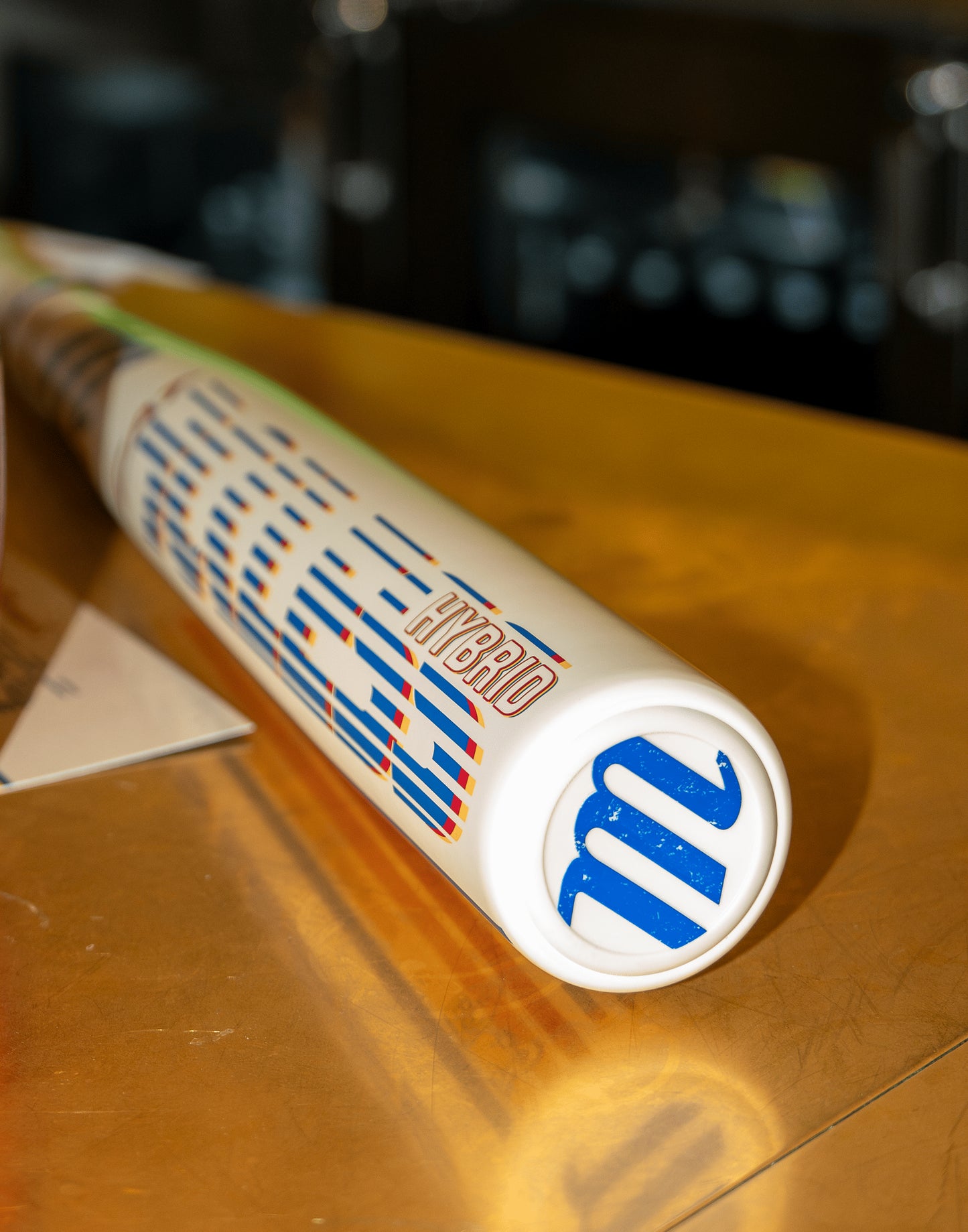 A close-up of the 2026 Marucci CATX RCKLESS Rewind Hybrid (-3) BBCOR Baseball Bat, featuring "HYBRID" and a blue M logo on the white barrel, rests on a golden-brown surface—ideal for power hitters.