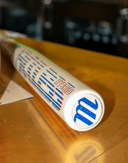 A close-up of the 2026 Marucci CATX RCKLESS Rewind Hybrid (-3) BBCOR Baseball Bat, featuring "HYBRID" and a blue M logo on the white barrel, rests on a golden-brown surface—ideal for power hitters.
