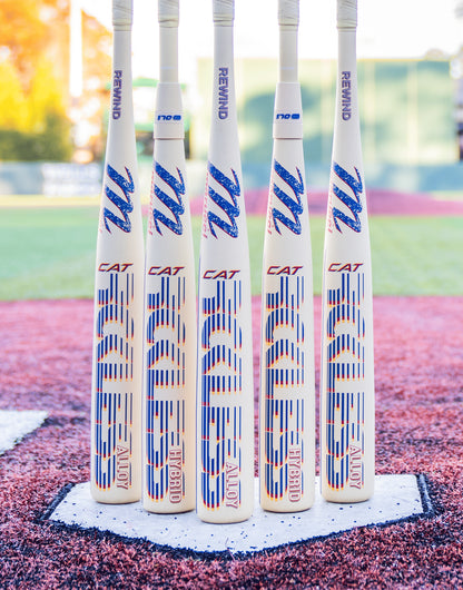 Five Marucci 2026 CATX RCKLESS Rewind Hybrid (-3) BBCOR bats with blue and red graphics stand upright on home plate, ready for a powerful swing, against a baseball field backdrop with a blurred green fence and trees.