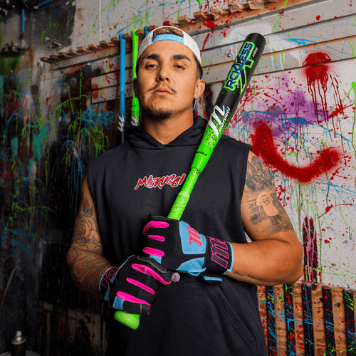 A man in a sleeveless hoodie, baseball gloves, and backward cap holds the 2026 Marucci CAT X RCKLESS Hybrid (-3) BBCOR Baseball Bat (MCBCRH DEMO), standing confidently before a wall splashed with colorful paint and graffiti.