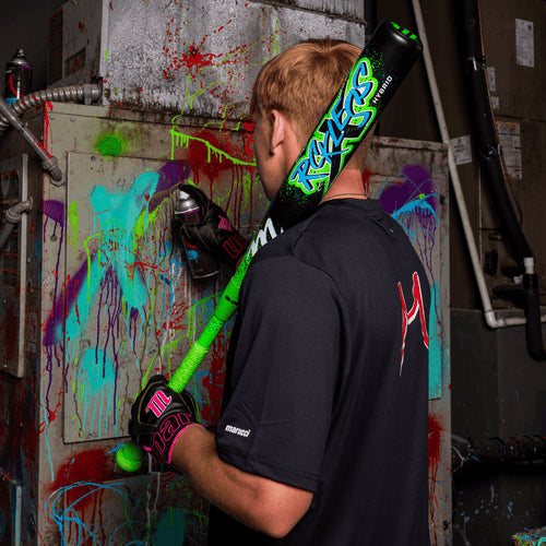 Wearing black gloves and a dark shirt, a person holds the Marucci CAT X RCKLESS Hybrid (-3) BBCOR Baseball Bat: MCBCRH (DEMO) over their shoulder, standing in front of a metal unit splattered with colorful paint.