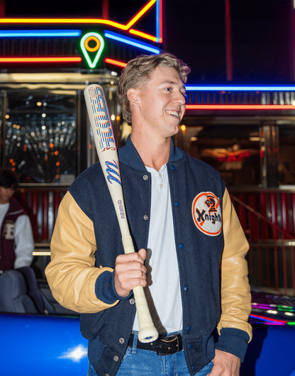 A young man in a varsity jacket smiles while holding the Marucci CATX RCKLESS Rewind (-3) BBCOR Baseball Bat (MCBCRRC) in front of a brightly lit neon diner at night.