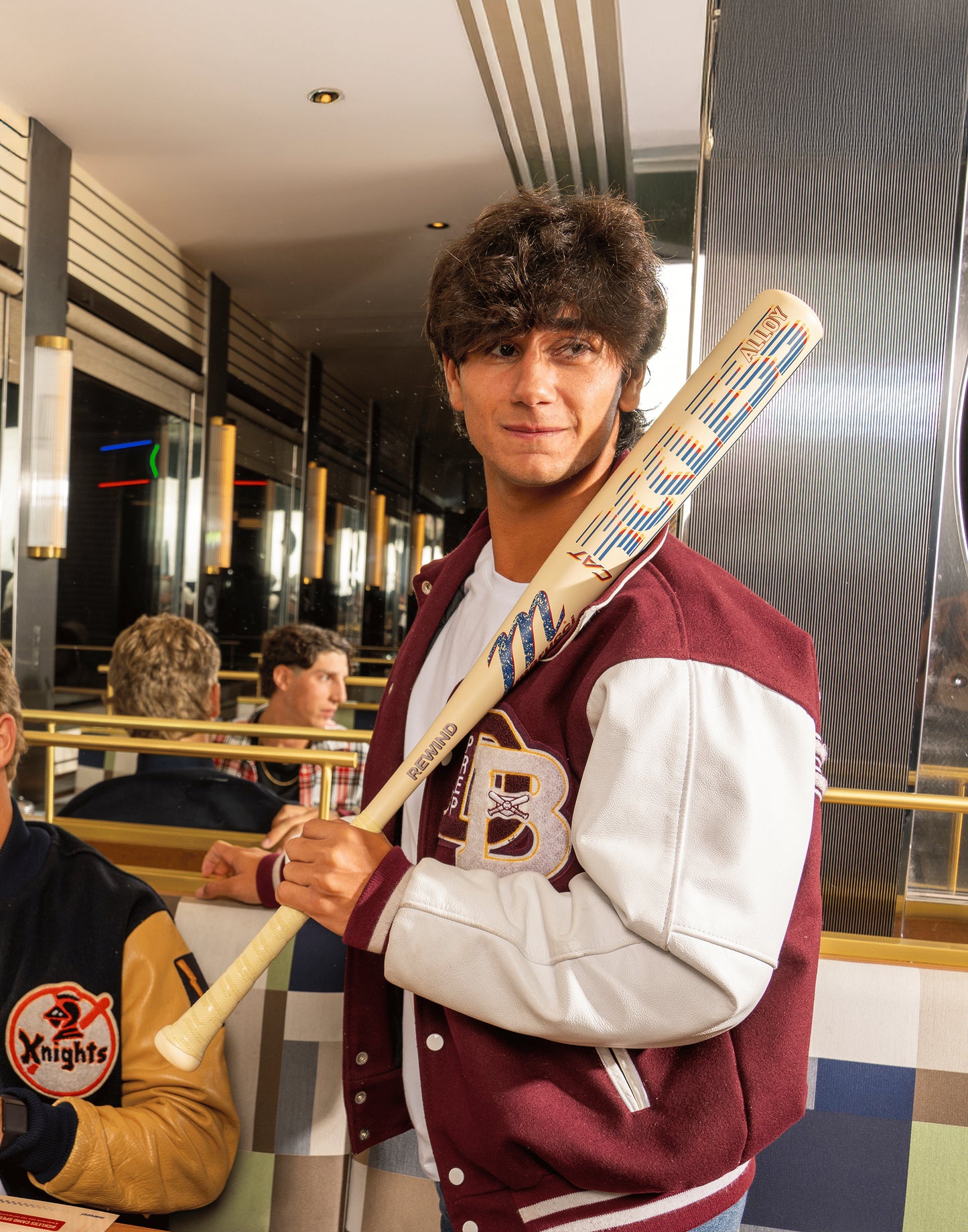 A young man in a maroon and white varsity jacket holds the Marucci 2026 CATX RCKLESS Rewind (-3) BBCOR Baseball Bat (MCBCRRC) over his shoulder, standing in a retro diner with mirrored walls and striped upholstery.