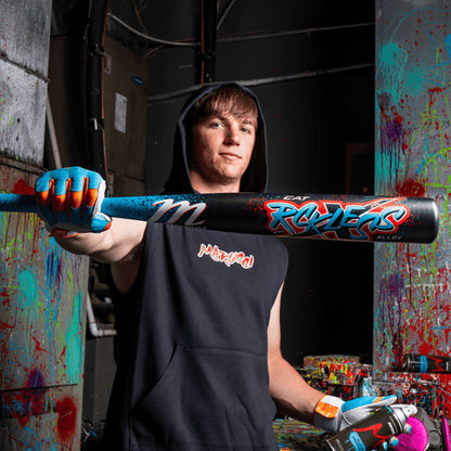 A young man in a sleeveless hoodie and colorful gloves points the 2026 Marucci CAT X RCKLESS (-3) BBCOR Baseball Bat (MCBCR) towards the camera, standing in a paint-splattered room with spray paint cans in his other hand.