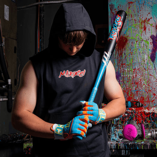 Wearing a sleeveless black hoodie and colorful gloves, someone looks down while holding a Marucci 2026 CAT X RCKLESS (-3) BBCOR Baseball Bat (MCBCR) in a graffiti-filled room with paint splatters on the walls and floor.