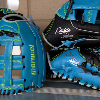 Close-up of the Marucci Caddo 11.5″ Baseball Glove (MFG3CD115-SB/BK) for youth, featuring a blue and black design with green and white stitching, crafted from full-grain cowhide leather by Marucci, displayed on a bench against a tan wall.