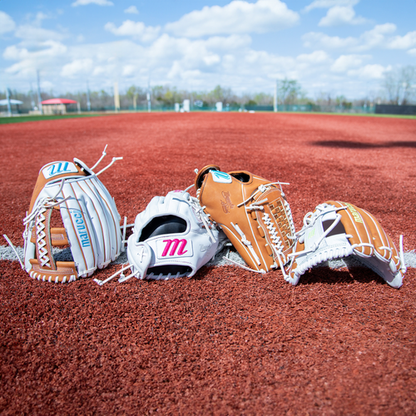 The Marucci Magnolia 39S2 13" Fastpitch First Base Mitts in tan, white, and brown are displayed side by side on red infield dirt beneath a bright, partly cloudy sky.