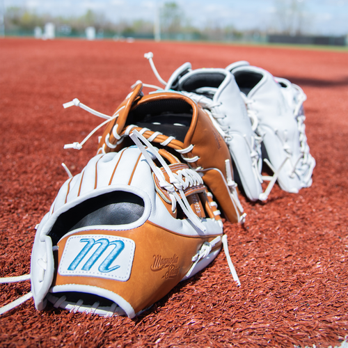 Three Marucci Magnolia 39S2 13" Fastpitch First Base Mitts, made with professional-grade rawhide laces and Japanese-tanned steerhide, are lined up on the red turf of a baseball field under a clear sky.