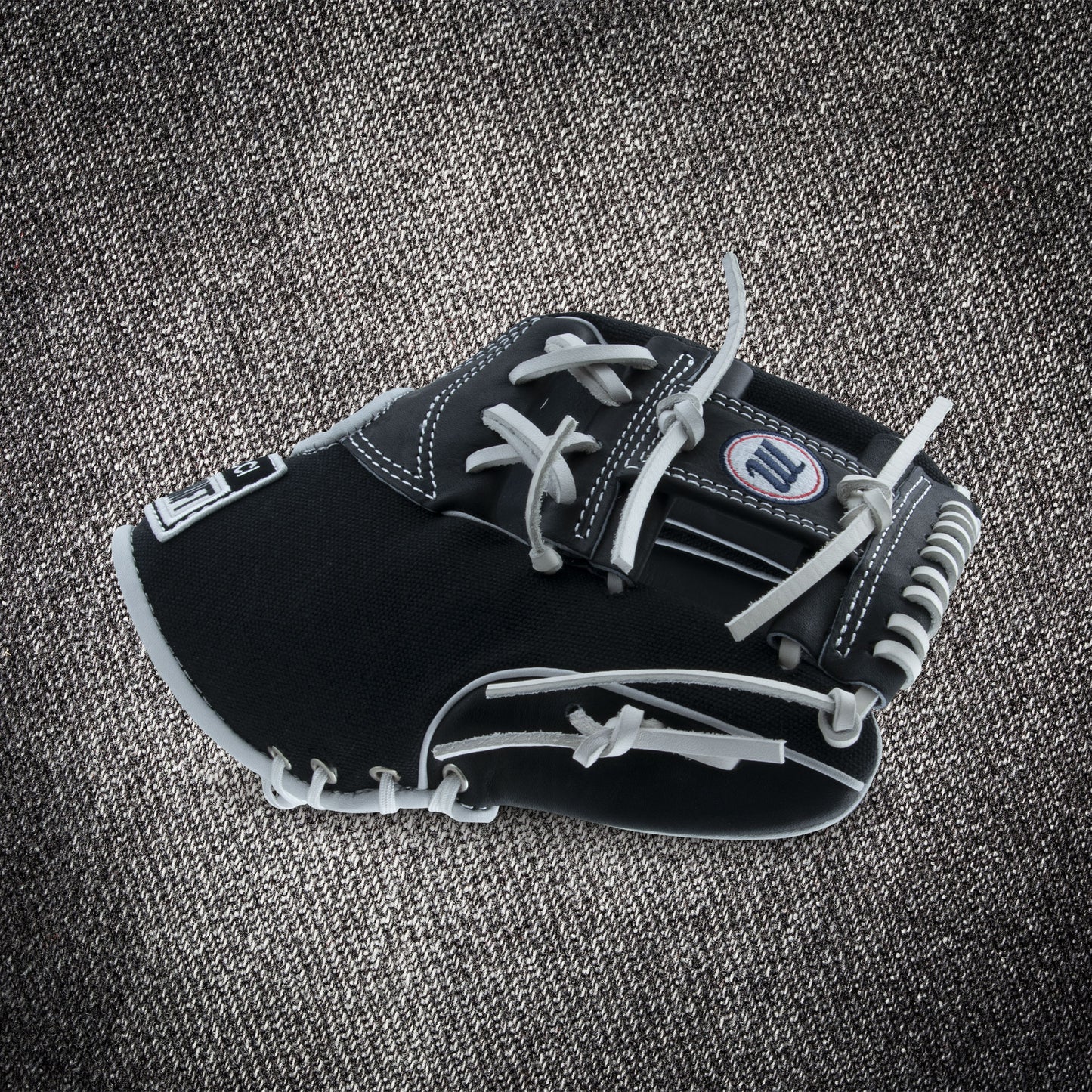 The Marucci NightShift CHUCK T 11.5" Baseball Glove (MFGNTSHFT-0103) in black and gray with white stitching and laces is shown on a textured gray fabric background.