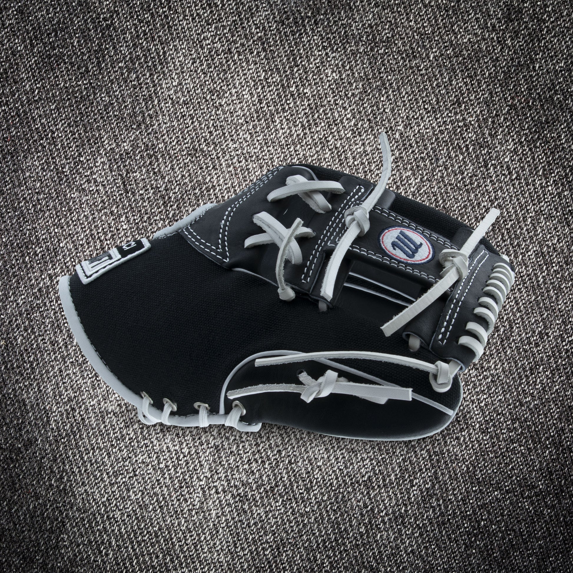 The Marucci NightShift CHUCK T 11.5" Baseball Glove (MFGNTSHFT-0103) in black and gray with white stitching and laces is shown on a textured gray fabric background.