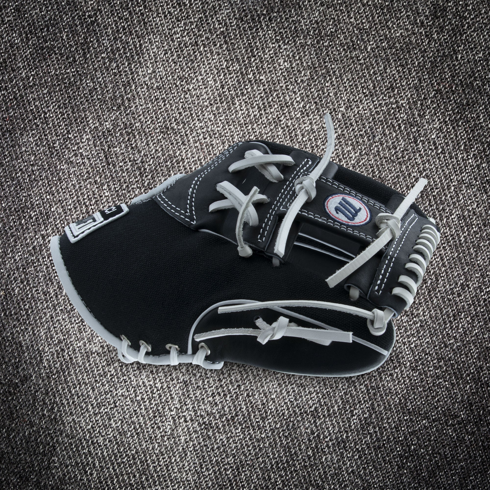 The Marucci NightShift CHUCK T 11.5" Baseball Glove (MFGNTSHFT-0103) in black and gray with white stitching and laces is shown on a textured gray fabric background.