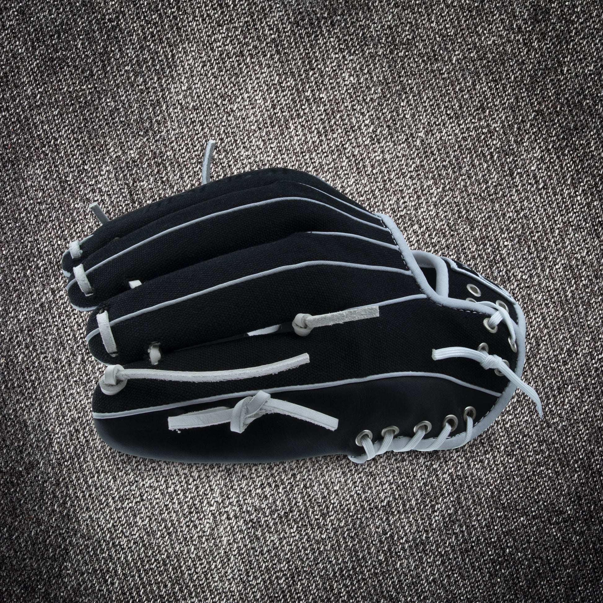 A Marucci NightShift CHUCK T 11.5" Baseball Glove (MFGNTSHFT-0103) from Marucci with white laces is shown closed on a textured gray surface.