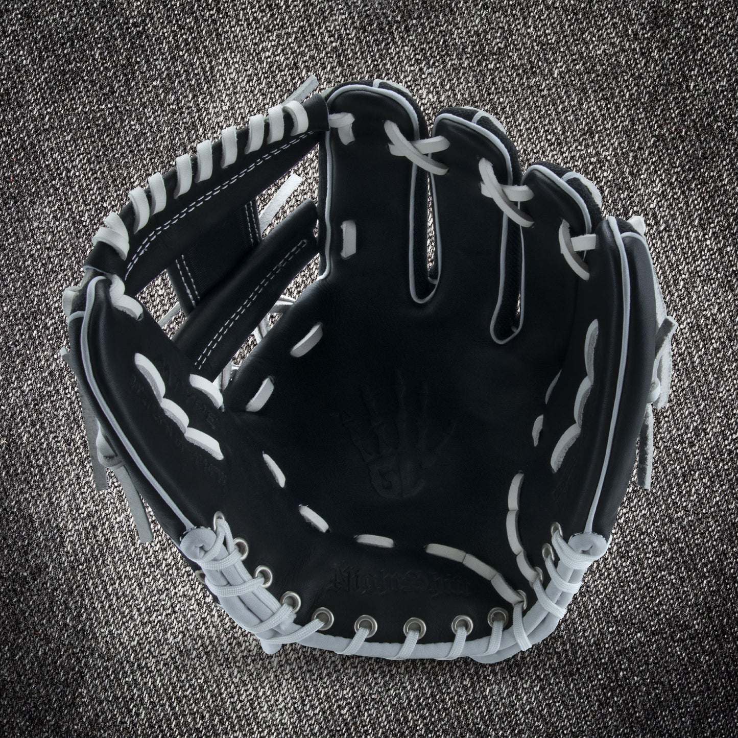 The Marucci NightShift CHUCK T 11.5" Baseball Glove (MFGNTSHFT-0103) in black with white lacing is shown open on a textured gray fabric background, highlighting the palm and finger slots.