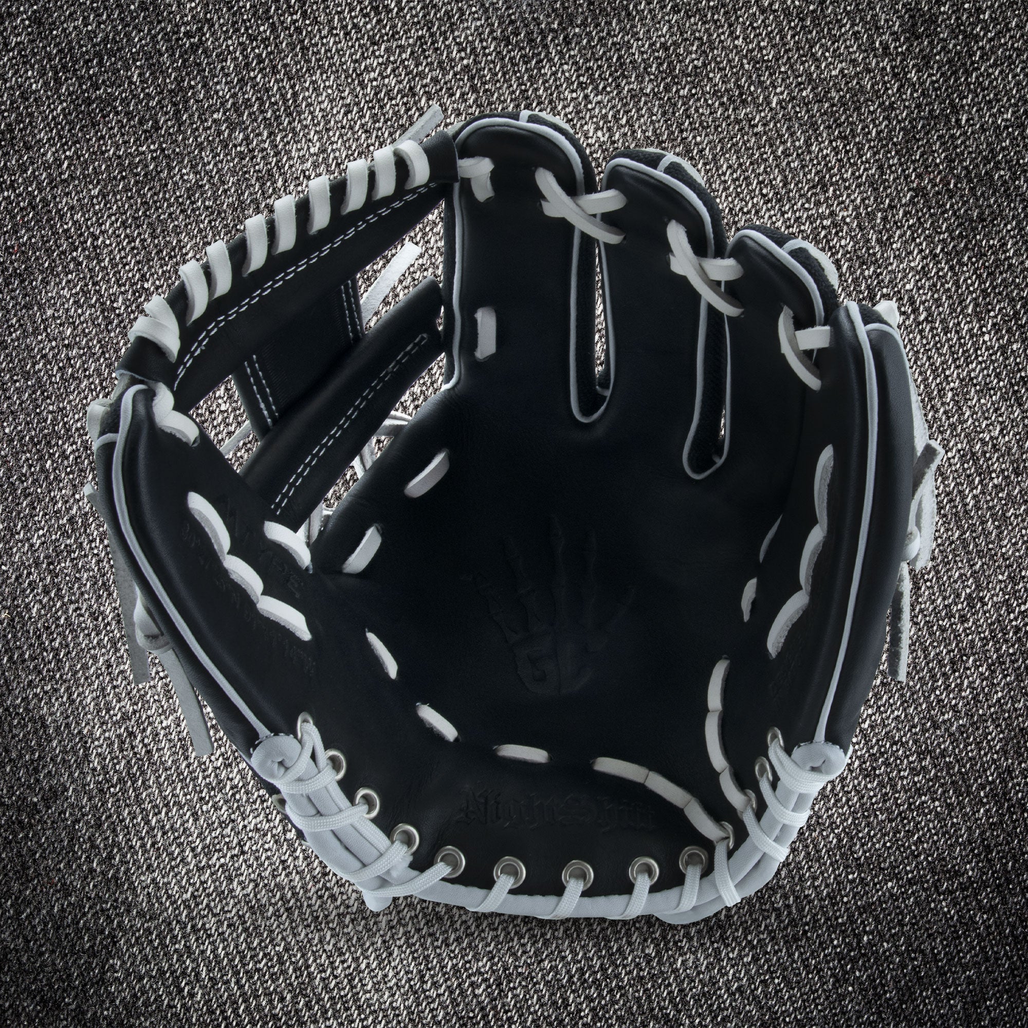 The Marucci NightShift CHUCK T 11.5" Baseball Glove (MFGNTSHFT-0103) in black with white lacing is shown open on a textured gray fabric background, highlighting the palm and finger slots.