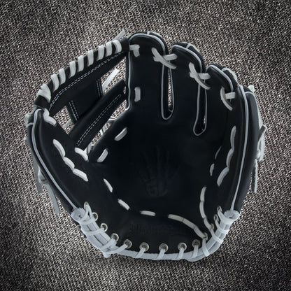 The Marucci NightShift CHUCK T 11.5" Baseball Glove (MFGNTSHFT-0103) in black with white lacing is shown open on a textured gray fabric background, highlighting the palm and finger slots.