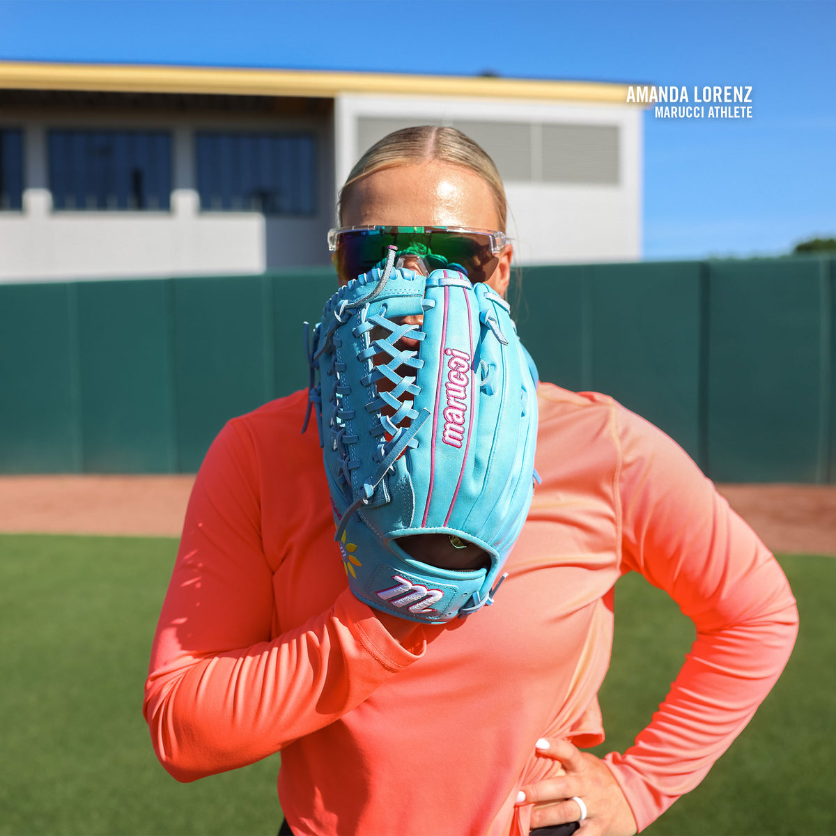 A softball player in sunglasses and a pink long-sleeve shirt holds a teal Marucci NightShift Lorenz 12.5″ Fastpitch Glove: MFGNTSHFT-0305 in front of her face on a sunny field. Text: Amanda Lorenz, Marucci Athlete.