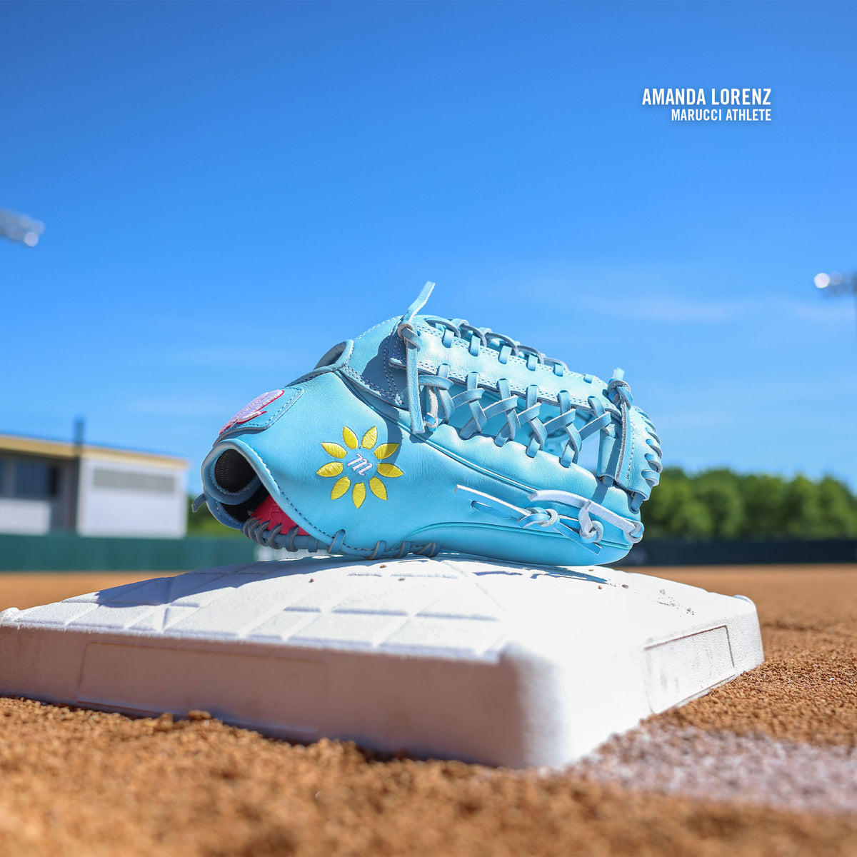 The Marucci NightShift Lorenz 12.5″ Fastpitch Glove: MFGNTSHFT-0305, in light blue with a yellow flower design, rests on a base on a dirt field beneath a clear blue sky. Text reads Amanda Lorenz Marucci Athlete.