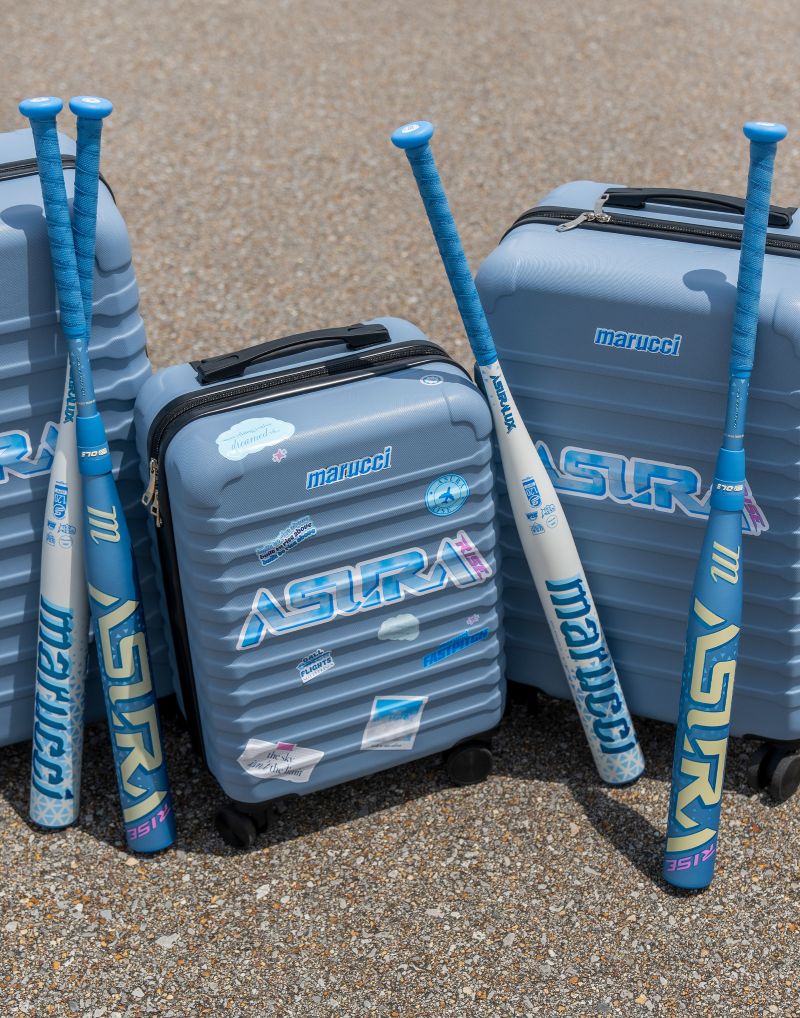 Three blue Marucci ASURA Rise (-11) Fastpitch Softball Bats, engineered with a wider sweet spot and an anti-vibration knob for enhanced comfort, lean against two blue Marucci suitcases decorated with travel stickers, all arranged on a gravel surface outdoors.
