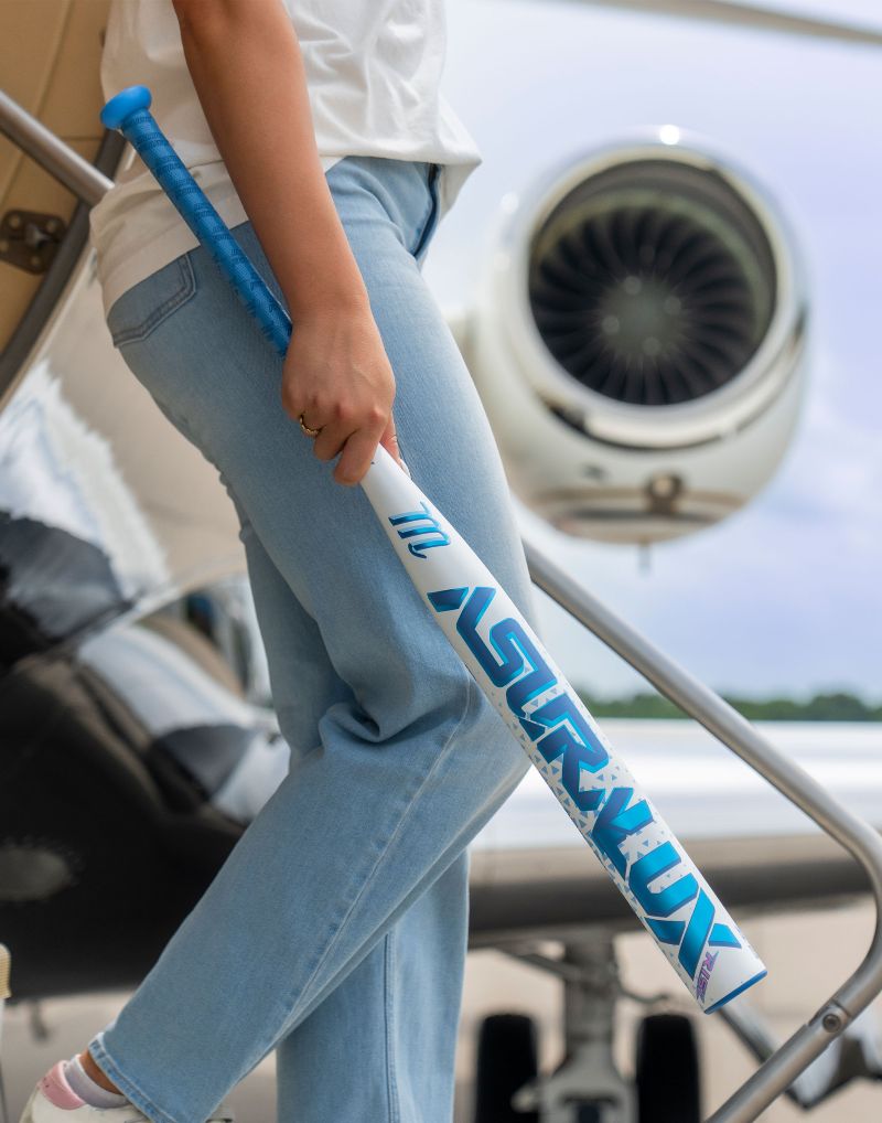 Wearing light blue jeans, a person stands on airplane stairs near a jet engine, holding a white and blue Marucci 2027 ASURA Lux Rise (-9) Fastpitch Softball Bat: MFPASLR9.