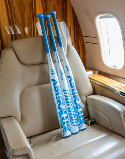 Three blue and white baseball bats, including the Marucci 2027 ASURA Lux Rise (-8) Fastpitch Softball Bat (MFPASLR8), are leaning against the back of a beige leather seat inside a private jet, next to a window.