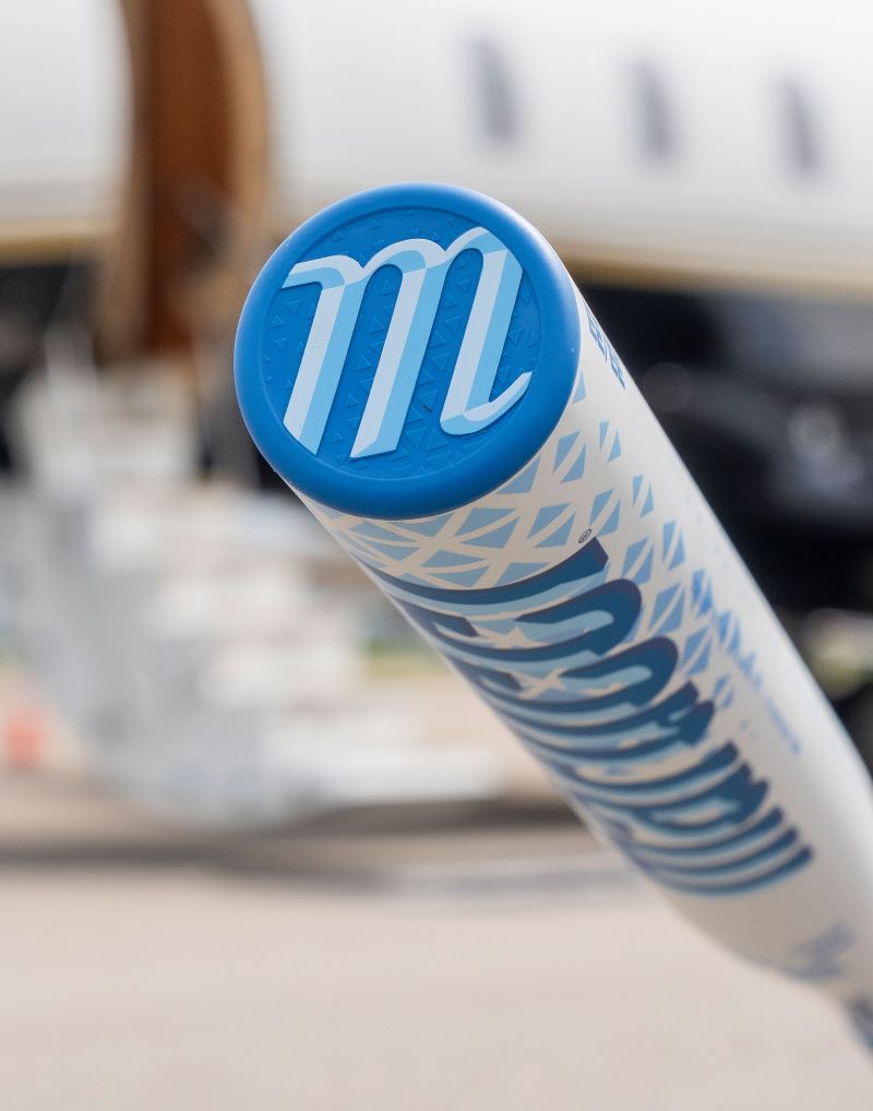 Close-up of the end of a Marucci 2027 ASURA Lux Rise (-11) Fastpitch Softball Bat (MFPASLR11), showcasing a stylized M on the AV Knob. The background is blurred, revealing stairs and part of an airplane.