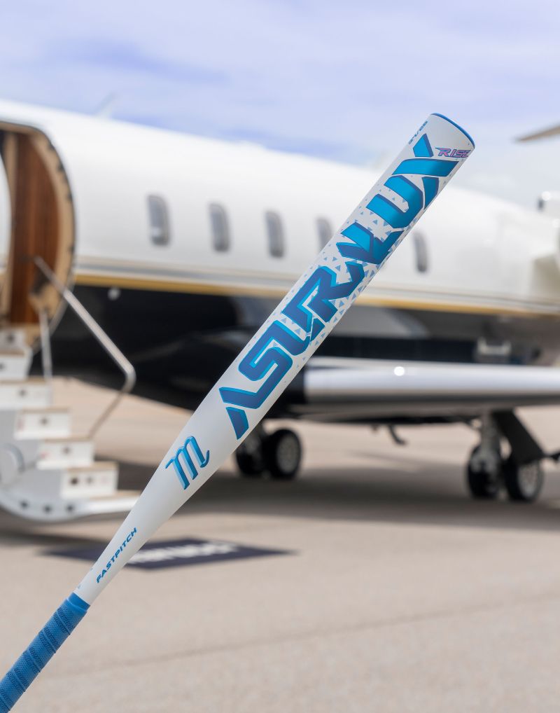 A Marucci 2027 ASURA Lux Rise (-11) Fastpitch Softball Bat (MFPASLR11), featuring Rise Sync Barrel Technology, is held upright in front of a private jet with stairs leading to its open door on an airport tarmac.