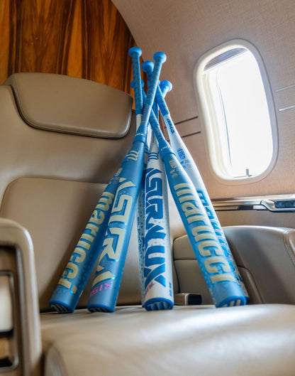 Four Marucci ASURA Rise (-11) Fastpitch Softball Bats, each featuring an anti-vibration knob for enhanced comfort, are arranged in a fan shape on a beige leather seat inside a private jet, illuminated by light streaming through a nearby window.
