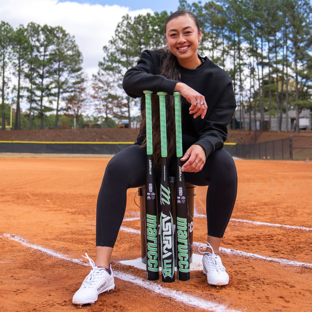 A woman in black athletic wear sits on a softball field stool, smiling as she holds three Marucci 2026 ASURA Lux (-10) Fastpitch Softball Bats (MFPASL10) upright. Trees and a fence are visible in the background.