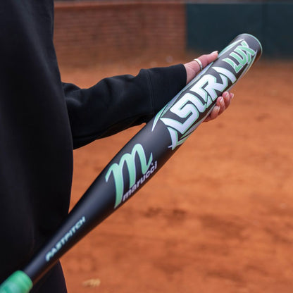 A person in a black long-sleeve shirt holds a Marucci 2026 ASURA Lux (-10) Fastpitch Softball Bat: MFPASL10 with a composite barrel over a reddish dirt field.