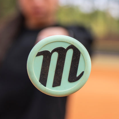 A close-up shot of a mint green circular object with Marucci’s bold black “M” logo, held by someone in dark clothing, mirrors the design of the 2026 Marucci ASURA Lux (-10) Fastpitch Softball Bat: MFPASL10.