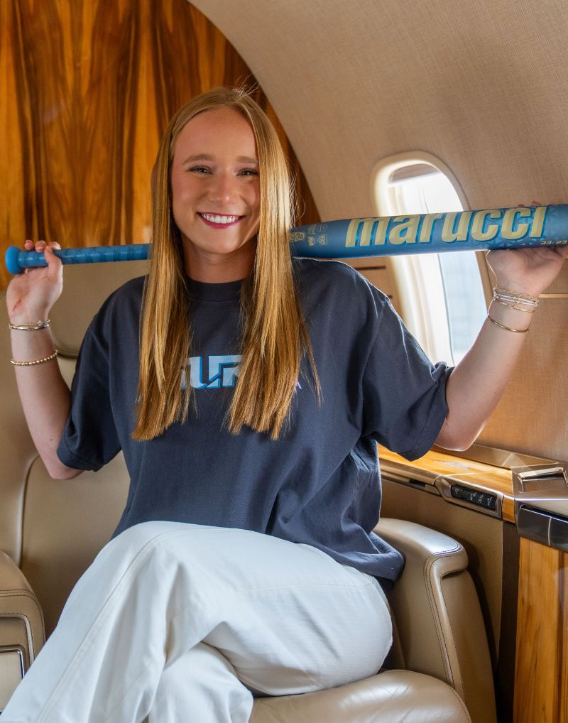 A smiling young woman sits in a private jet holding a blue Marucci 2027 ASURA Rise (-11) Fastpitch Softball Bat (MFPASR11) with an anti-vibration knob across her shoulders, wearing a dark t-shirt and white pants.
