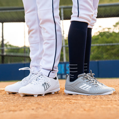 Two people in white baseball pants and navy socks stand on a baseball field, each wearing different pairs of Marucci FLT Youth Molded Cleats (MFWFLMY) in gray and white, facing forward on the dirt infield.
