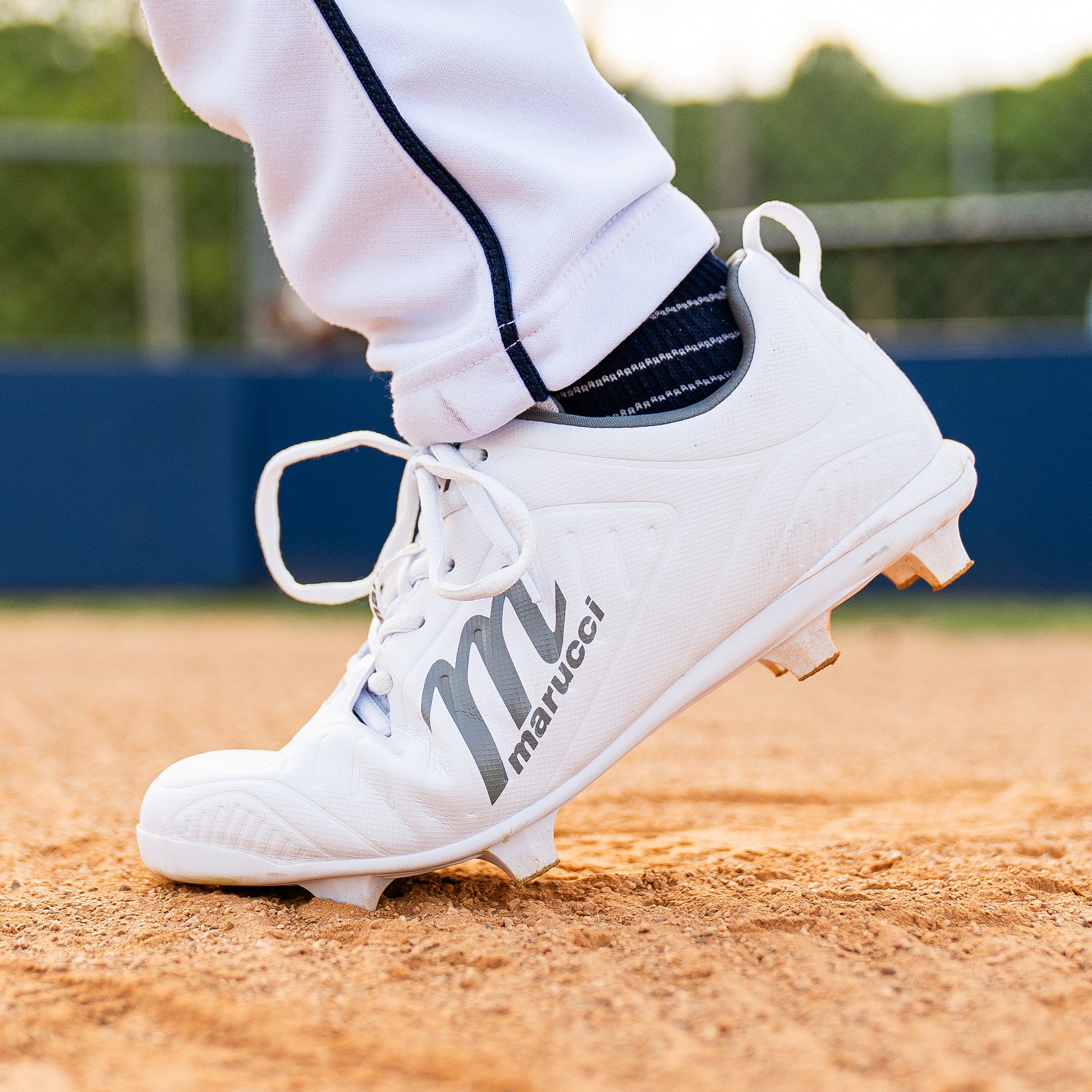 A close-up of a person wearing Marucci FLT Youth Molded Cleats (MFWFLMY) standing on a dirt baseball field, with part of a white uniform pant leg visible.