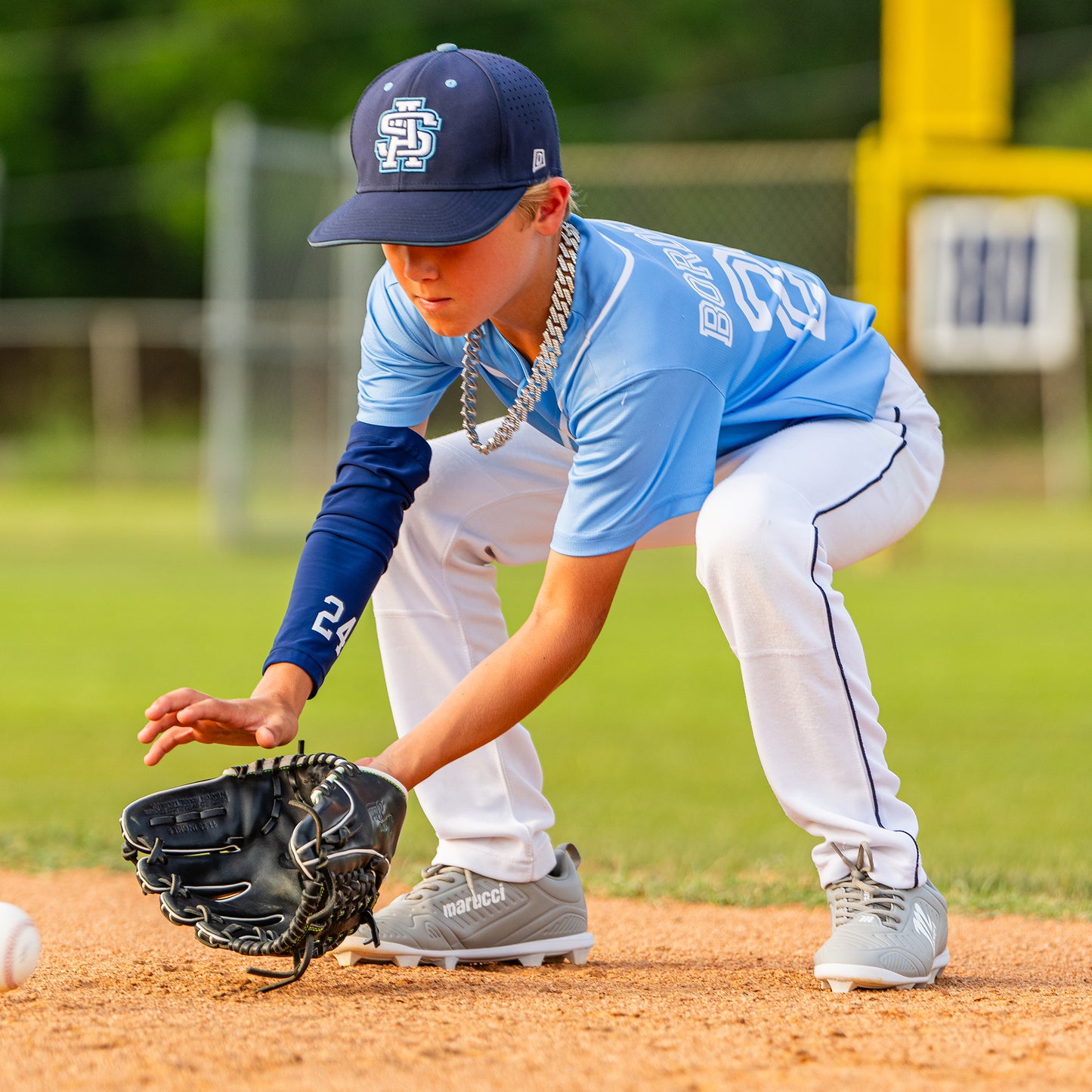 A young baseball player in a blue uniform and cap bends down with a glove to field a ground ball on the baseball field, his Marucci FLT Youth Molded Cleats: MFWFLMY from Marucci digging into the dirt for quick, agile movement.