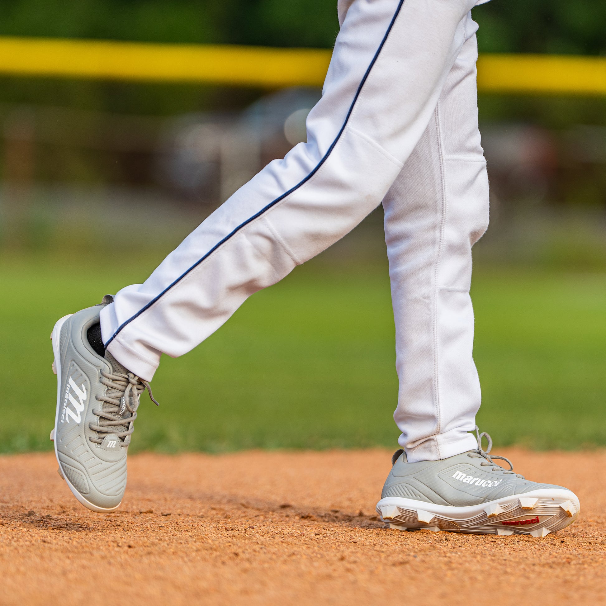Wearing white baseball pants and Marucci FLT Youth Molded Cleats (MFWFLMY), a child stands on a baseball field, one foot lifted while the other presses into the dirt. Behind them, green grass and a yellow fence complete the scene.