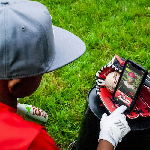 A young baseball player in a gray cap and red shirt, sitting on a bucket on green grass, uses a smartphone displaying a baseball app beside his Marucci 2024 CATX SMART (-5) USSSA Baseball Bat, glove, and ball.