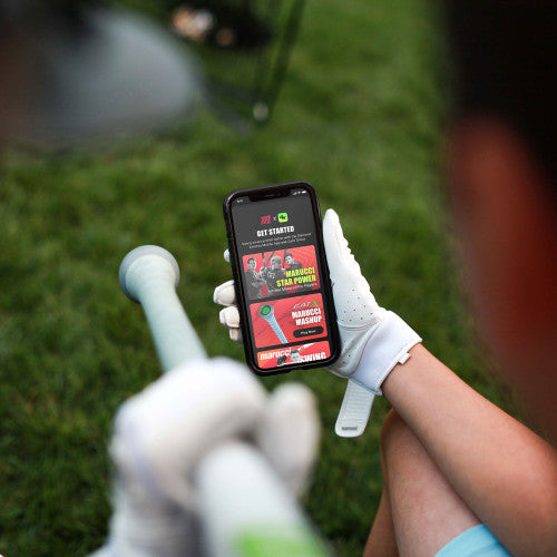 A person wearing a white glove holds a baseball bat and looks at a smartphone displaying a colorful app interface, possibly related to baseball training or equipment.