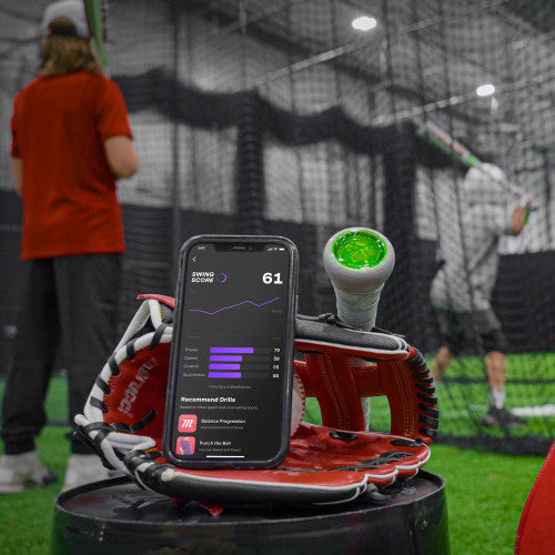 A smartphone displaying a swing score and stats rests on a baseball glove, with a bat and green sensor nearby. In the background, people practice baseball in an indoor batting cage.