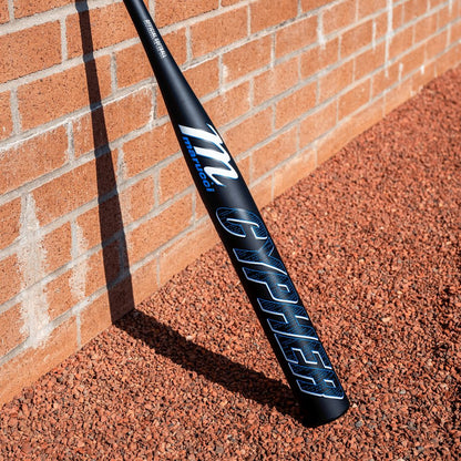 A black 2025 Marucci Cypher 12.25" BH Load NSA USSSA Slowpitch Softball Bat (MSPC1BHU), with blue and silver lettering and a Dynamic Performance Optimized Barrel, leans against a brick wall on reddish gravel.