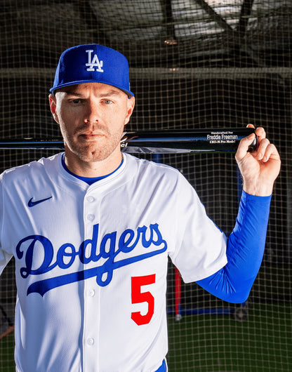 A baseball player in a Dodgers uniform and cap poses holding a black Marucci FREEMAN5 Freddie Freeman Pro Exclusive Maple Wood Bat (MVE4FREEMAN5) over his shoulders, with the number 5 visible on his jersey. A netted backdrop and indoor lighting are seen in the background.