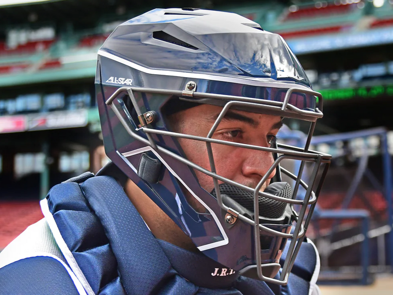 A baseball catcher wearing a navy blue helmet and protective face mask is focused on the game, with stadium seats and part of a batting cage visible in the background.
