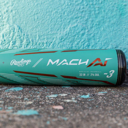 A used 2024 Rawlings Mach AI (-3) BBCOR Baseball Bat (RBB4MC3) in teal with red and white lettering rests on worn concrete against a turquoise wall. The alloy bat features a 33" length, 2 5/8" barrel, and -3 drop.