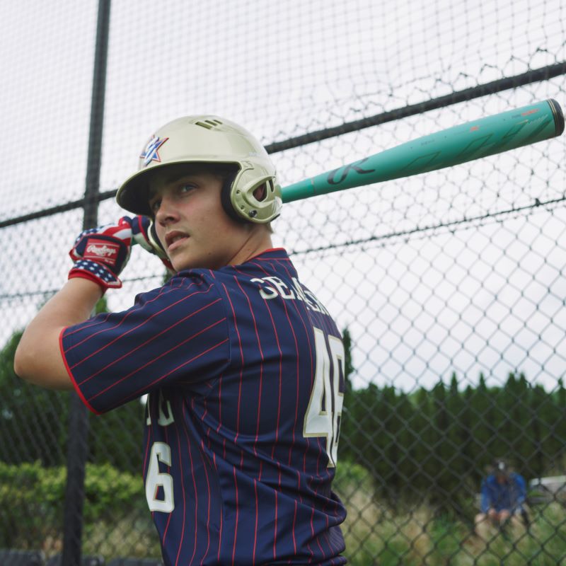 A baseball player in a striped navy jersey and helmet holds a used 2024 Rawlings Mach AI (-3) BBCOR Baseball Bat: RBB4MC3 by Rawlings over his shoulder, standing ready to hit near a chain-link fence on a cloudy day.