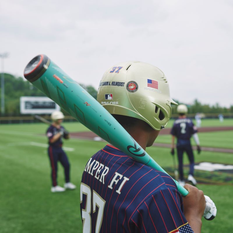 A youth player in jersey #37 stands on the field with a Rawlings 2024 Mach AI (-3) BBCOR Baseball Bat (RBB4MC3, USED) resting on his shoulder; other players and a scoreboard appear in the background.